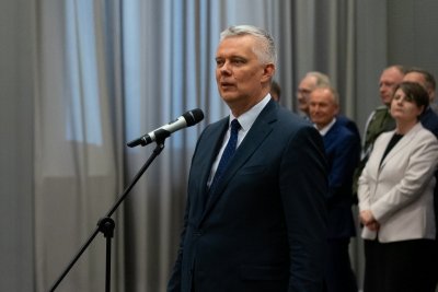 Mężczyzna w garniturze w trakcie przemowy. Za nim sylwetki elegancko ubranych ludzi.