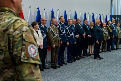 Ludzie w garniturach oraz mundurach stoją w szeregu, w tle flagi Polski i Unii Europejskiej.