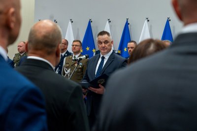 Mężczyzna w garniturze w trakcie przemowy, na pierwszym planie plecy mężczyzn w garniturach, w tle mężczyźni ubrani w mundury.