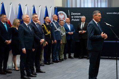 Mężczyzna w garniturze w trakcie przemówienia, za nim sylwetki osób w garniturach i mundurach.