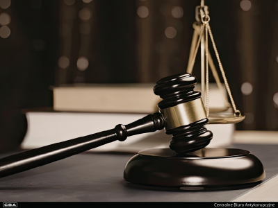 Drewniany młotek sędziowski oparty o podstawkę oraz mosiężną wagę Temidy w tle. Obok leżą grube księgi. Na dole na czarnym pasku znajduje się logo CBA o napis Centralne Biuro Antykorupcyjne.