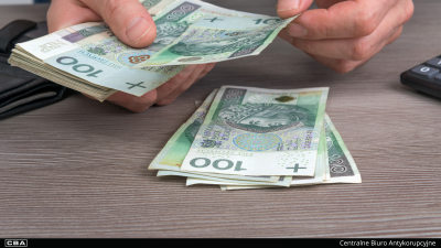 Męskie dłonie odliczające pliki polskich banknotów o nominale 100 złotych na drewnianym blacie biurka. Po lewej stronie widoczny jest fragment czarnego portfela, a po prawej – róg kalkulatora. Na dole na czarnym pasku znajduje się logotyp oraz napis Centralne Biuro Antykorupcyjne.
