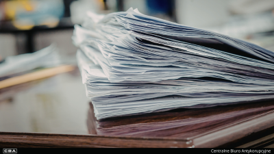 Stos białych dokumentów i kartek papieru, ułożonych niesymetrycznie na ciemnym, błyszczącym blacie biurka. W lewym dolnym rogu widoczne jest logo CBA, natomiast w prawym dolnym rogu umieszczono podpis „Centralne Biuro Antykorupcyjne”