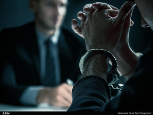 An officer writing a note and a man sitting with the handcuffs on.