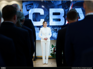 Ms Agnieszka Kwiatkowska-Gurdak, the Head of the Central Anti-Corruption Bureau stands in front of the people in suits. A blue caption reading ‘CBA’ in the background.