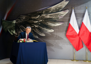 Mr Tomasz Siemoniak, Minister of the Interior and Administration, Coordinator of Special Services sits at the table and writes. Two Polish flags and eagle wing figure in the background.