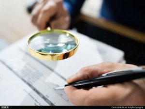 A man holds a pen in the left hand and reads a document through a magnifier held in the right hand.