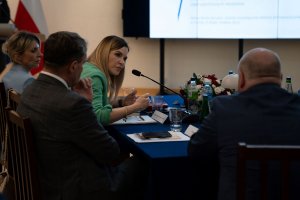 Head of the CBA, Ms Agnieszka Kwiatkowska-Gurdak sits at a table with the attendees and speaks through a microphone.