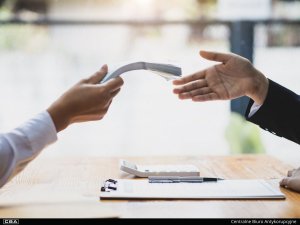 Two people sit at a table. One hands over a wad of money. The other extends his hand.