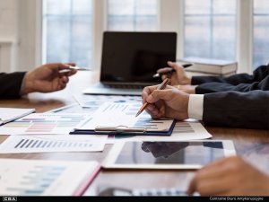 A group of people sit at a table and hold pens. A laptop, tablet and documents on the table.