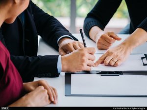 Three people sit at a table. A woman points to a place on a document and a man signs. The other woman sits next to him.