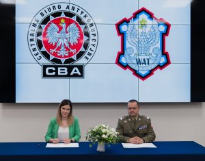 A photo of the Commandant-Rector of the WAT, Brigadier General Prof. Przemysław Wachulak and the Head of the CBA, Agnieszka Kwiatkowska-Gurdak. They hold files with the WAT and CBA emblems. CBA and WAT emblems in the background.