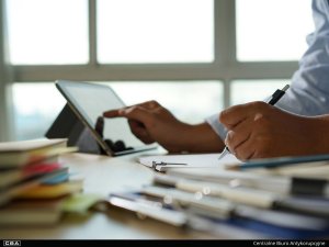 A man sits at the table and writes. He points at a tablet.