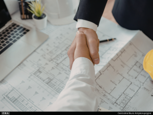 Shaking hands in the foreground, development plans on the table in the background.