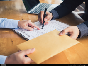 A close up of two men sitting at the desk, handing each other an envelope.