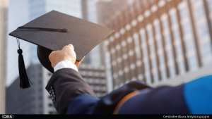 A close-up of a man's hand with a graduation cap, a building in the background.